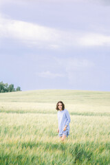 A young girl in a blue dress stands in a field with tall grass, against the background of a clear...