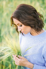 A girl with brown hair in a light blue sweater holds a wheat ear, looking at it thoughtfully. The...