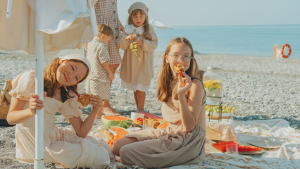 Group of happy kids enjoying a summer picnic on the beach with watermelon, lemonade, grapes, and...