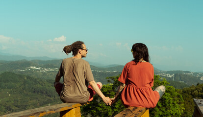 Young couple holding hands, sitting on wooden railing and enjoying scenic mountain view together...