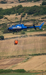 Firefighting helicopter flies with water bucket above wind turbine and forested hills