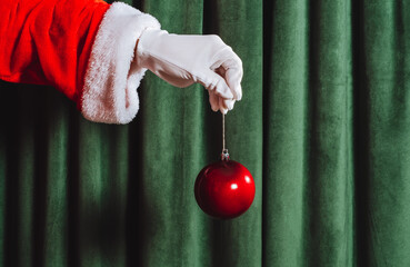 Santa's gloved hand holding a red Christmas ornament on green velvet background. Minimalist festive holiday concept.