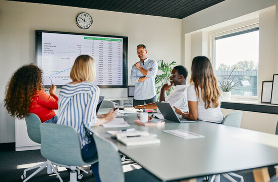 Team Presentation in Modern Office With Diverse Group Engaged in Discussion. A businessman with gray hair stands smiling at the whiteboard and presents a topic to his younger diverse colleagues