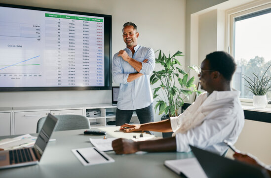 Business Professionals Engaged in a Lively Office Presentation Meeting. A businessman with gray hair stands smiling at the whiteboard and presents a topic to his younger diverse colleagues