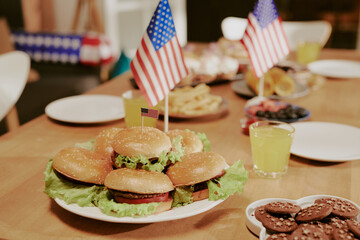 Plate of hamburgers with lettuce arranged on wooden table, small American flags inserted in buns, background showing plates of cookies, fruit, and glasses of fizzy drink 