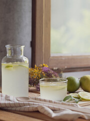 Refreshing lemonade along with lemons and limes on a wooden table next to a country house window in summer