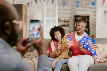 Black woman and Black child sitting on sofa holding American flags and smiling while man taking photo with smartphone during patriotic celebration at home