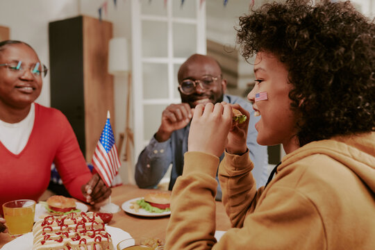 Teenage Black boy with American flag face paint eating sandwich while sitting at table with mom and dad holding small American flag