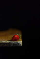 The image shows a single strawberry lying on the edge of a wooden surface. The background is black, which creates a contrast with the bright red berry illuminated by the light.
