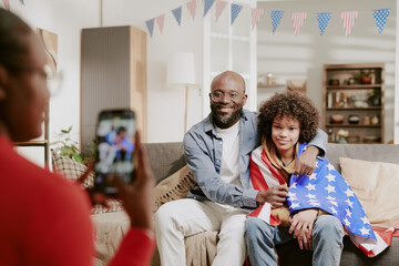 African American father sitting on sofa embracing teenager wrapped in American flag while woman photographing them with smartphone during family gathering in living room