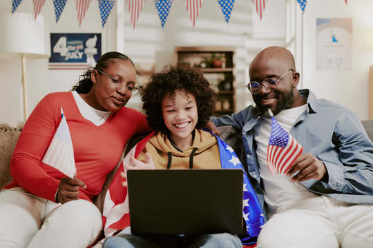 Family sitting together on sofa smiling and holding American flags while using laptop