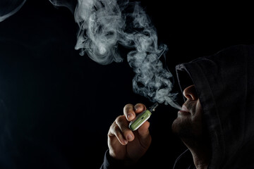 Hooded Man Vaping with Electronic Cigarette Exhaling Dense Vapor on Black Background, Modern...