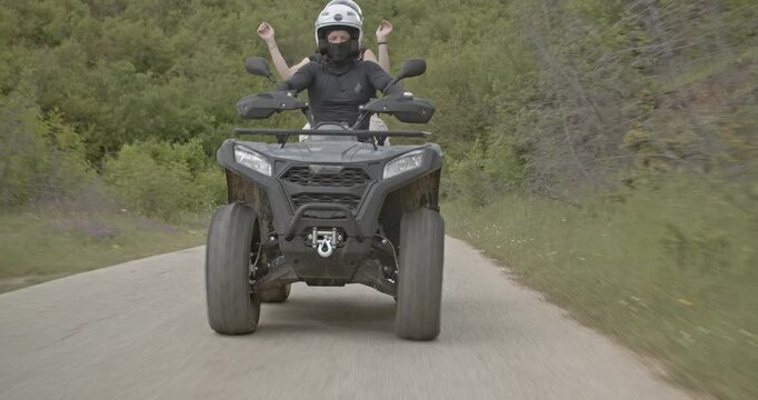 Two friends enjoy riding on a four wheeler ATV, putting their arms up in the air and smiling as they drive through a forest.