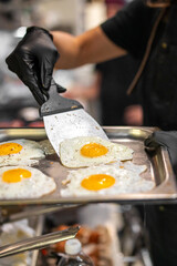 Chef in black gloves serving sunny-side-up eggs with runny yolks and pepper seasoning from a metal tray in a professional kitchen setting.