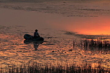 A man catches fish on an inflatable boat on the river in the evening with the setting sun. A...