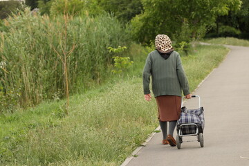 An elderly woman walks along a green avenue with a product cart. The difficult fate of people of declining age. Care for the elderly. Grandma's loneliness