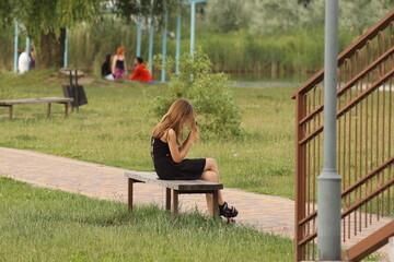 A teenage girl is sitting on a bench in a park in a black dress with a cell phone. Fashion of modern youth. Young girl relaxes in nature in a sunny park.