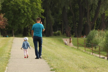 A father and his little child are walking along the alley of a summer park. A man and a girl are walking in nature. Relaxation in nature for the harmonious development of a child. Relationship between