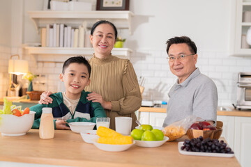 cheerful Asian family shares heartwarming moments as grandparents enjoy breakfast conversation with their grandson at home. The grandmother and young boy wash fresh fruits together in the open kitchen