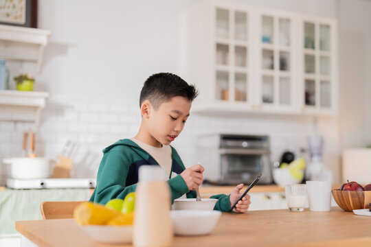 teenager enjoys a casual morning at home, multitasking with a smartphone while having breakfast in the pantry. The young student studies online, representing the modern lifestyle of tech-savvy kids ba