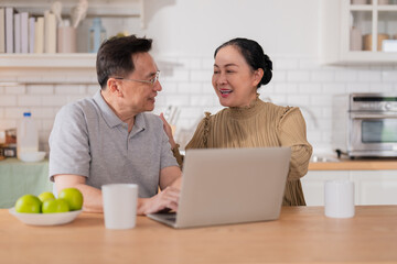 Asian senior couple sitting at the kitchen dining table, smiling warmly as they video call their grandson to celebrate his birthday online. Embracing technology, the grandparents use a digital tablet