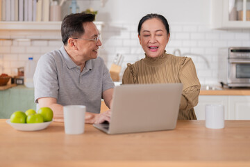 Asian senior couple sitting at the kitchen dining table, smiling warmly as they video call their grandson to celebrate his birthday online. Embracing technology, the grandparents use a digital tablet 
