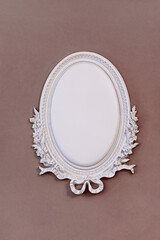 Ornate white oval frame with intricate floral designs placed against a soft brown wall, providing a...