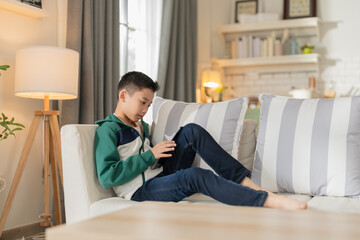 smart and cheerful Asian boy studies at home, typing on his computer and using a smartphone to...