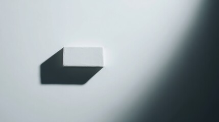 Minimalist composition featuring a small rectangular white object on a light-toned surface, casting a distinct dark shadow to the left, with a gradient background transitioning from bright to dark.