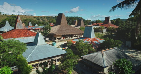 Aerial view of a village in Sumba, Indonesia, showing traditional houses with conical roofs made of straw and metal sheets, surrounded by lush vegetation under a clear blue sky