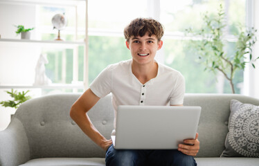 Handsome serious teenage boy sitting at home on the couch typing on laptop