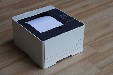 A compact printer is on the floor which is a wooden surface. A sheet of paper peeking out from its top output slot.