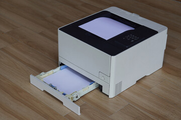 A black and white laser printer sitting on a wooden floor. A stack of white paper is visible in the main paper output tray on top of the printer and the lower paper input tray is pulled out.