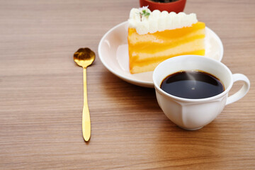 Hot black coffee in a white cup, with a spoon and a piece of cake beside it, all placed on a large wooden table.