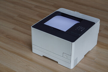 A laser printer positioned on a wooden floor with a sheet of white paper visibly exiting its top slot. its input tray is not extended or visible in the frame.