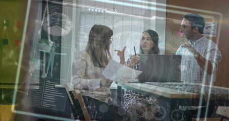 Team in business attire discussing data overlay around high table in office, with laptop, docs, pen