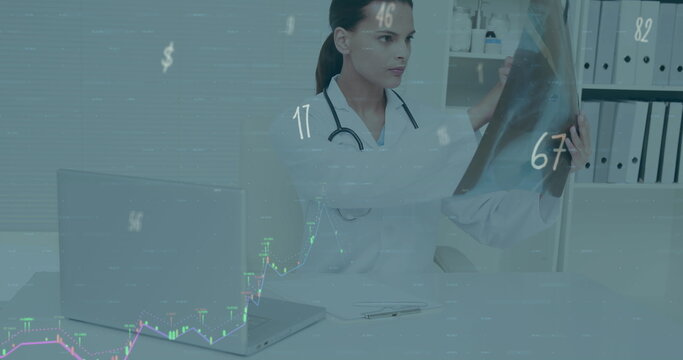 Female doctor wearing coat and stethoscope examining X-ray film in clinic, with laptop, charts
