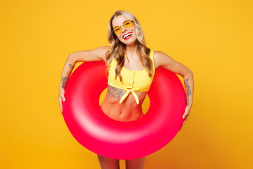 Young smiling fun beautiful sexy woman wear bikini swimsuit sunglasses rest near hotel pool hold pink rubber ring look camera isolated on plain yellow background. Summer vacation sea sun tan concept.