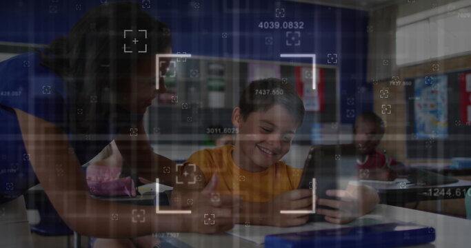 Guiding female teacher leaning over desk in school classroom, assisting smiling boy using tablet - Powered by Adobe