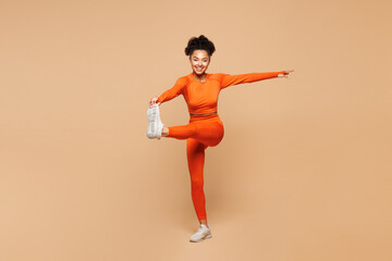 Full body young fitness trainer instructor sporty woman sportsman she wear orange clothes spend time in gym raise up leg touch foot isolated on plain beige background. Workout sport fit abs concept.