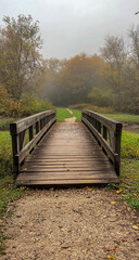 Fototapeta premium A rustic wooden bridge spans a quiet river, shrouded in thick fog amidst vibrant autumn foliage, creating a serene and mysterious atmosphere.