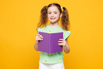 Little smiling intelligent cheery fun cute kid child girl 8-9 years old wears green t-shirt read purple book look camera isolated on plain yellow background Childhood kids education lifestyle concept