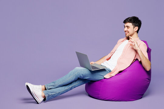 Full body young IT man he wearing pink shirt t-shirt casual clothes sit in bag chair hold use work on laptop pc computer waving hand talk speak isolated on plain purple background. Lifestyle concept. - Powered by Adobe