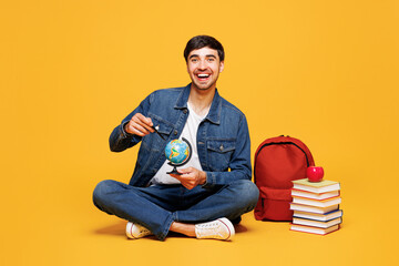 Full body young boy student he wear denim casual clothes sitting near backpack bag books hold in hand spin Earth map globe isolated on plain yellow background. High school university college concept.