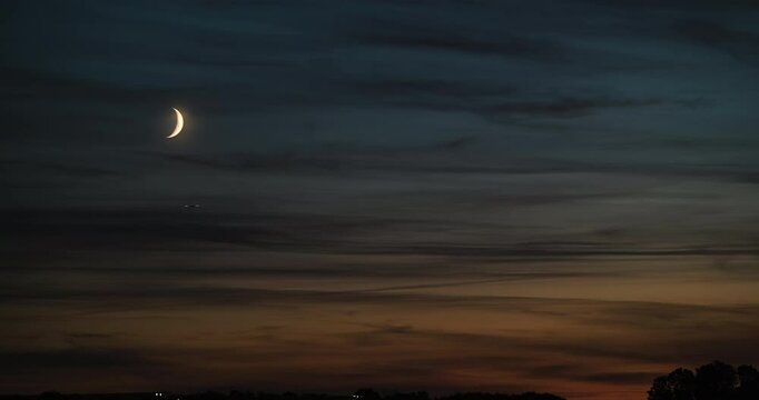 Timelapse of the nightfall scenery with dark orange and blue sky after sunset with wispy clouds and crescent moon slowly sinking toward the horizon