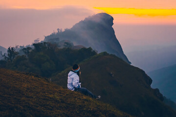 Fototapeta premium Photo of a woman sitting on a peak of the mountain during early sunrise. The sky is clear with warm tones of orange, yellow, and pink. Feeling fresh and free. The idea for trekking wallpaper.