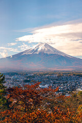 Obraz premium Cityscape views of the city near the volcano in the autumn from the Chureito Pagoda, Japan. There is Mount Fuji with snow on the top with a clear blue sky. There are colorful houses and buildings.