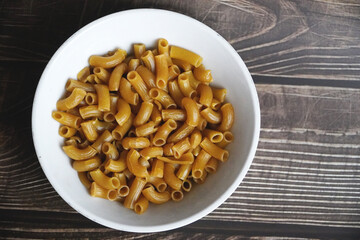 close up of freshly raw elbow pasta in a white ceramic bowl placed on a wooden table
