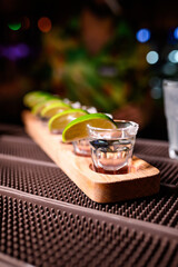 A row of tequila shots garnished with lime wedges and salt rims, arranged on a wooden tray on a bar counter. Party or bar atmosphere with focus on the front glass