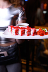 A waitress in a shiny silver top serves a plate of California rolls covered in red tobiko. Wasabi, pickled ginger, and soy sauce are also on the plate. Nightlife restaurant or club setting